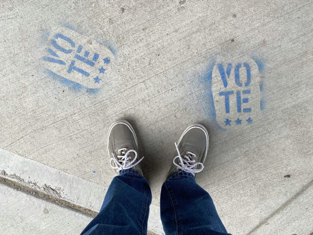 A person standing over two spray-painted logos that say "VOTE" on the sidewalk